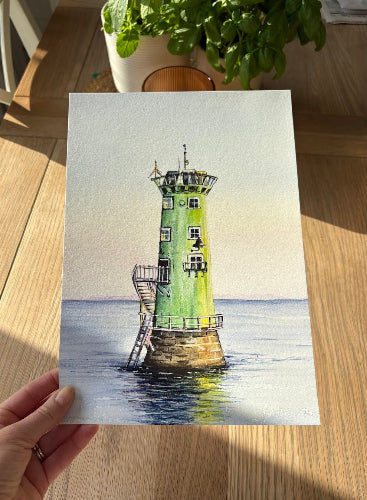 Hand holding a painting of a lighthouse on a wooden surface with a plant in the background.