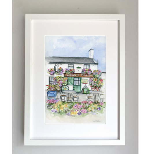 Framed artwork of the outside of the quaint pub Johnnie Foxes with flowering muti colored hanging baskets and outdoor seating on a gray wall.